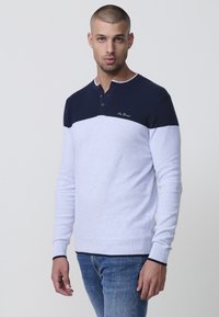 Šviesiai pilka ir tamsiai mėlyna ilga rankovė megztinis su henley apykakle, tekstūruota viršutine dalimi ir raštuotais rankogaliais. Yra trys mygtukai ir siuvinėtas logotipas.