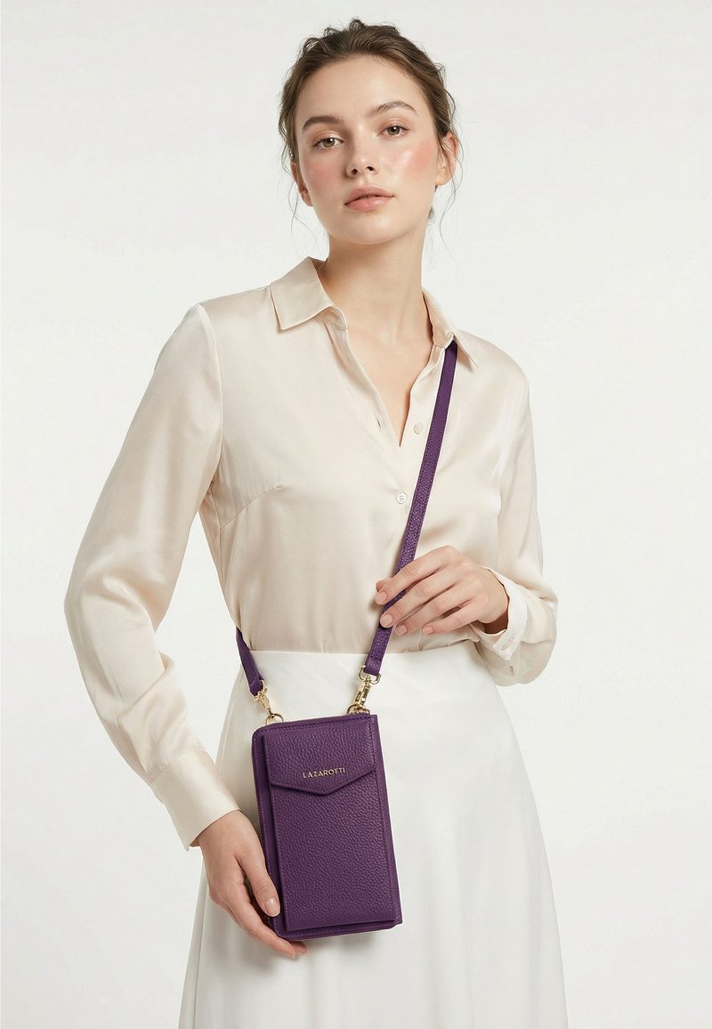Mujer vestida con una blusa y falda crema sostiene un pequeño bolso bandolera morado con herrajes dorados sobre un fondo blanco liso.
