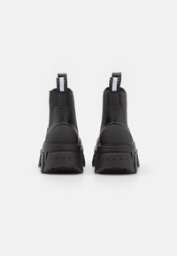 Pár černých chelsea bot s texturovaným podpatkem, silnou podrážkou a značkovým poutkem a patou s nápisem "TOMMY" na bílém pozadí.