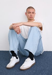 Hvid kortærmet skjorte, lyseblå baggy jeans og hvide sneakers med grå detaljer og brune såler. Siddende stilling mod en hvid væg.
