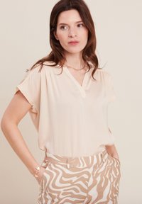 Beige V-Ausschnitt-Bluse mit kurzen Ärmeln und dekorativen Nähten an den Schultern, kombiniert mit sandfarbigen und weißen gestreiften Hosen. Seidige Textur.