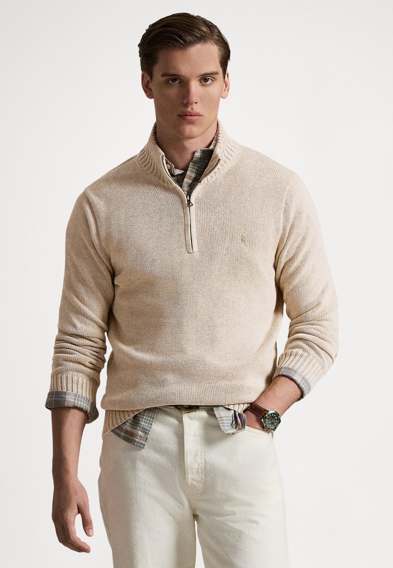 Beige geribbelde gebreide trui met een kwartsluiting en een hoge hals, gedragen over een geruit overhemd, gecombineerd met crèmekleurige broek en een polshorloge.