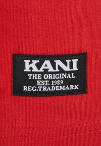 Sarkans audums ar teksturētu virsmu, uz kura ir melns uzlīmes marķējums ar tekstu "KANI THE ORIGINAL EST. 1989 REG. TRADEMARK" baltā krāsā.