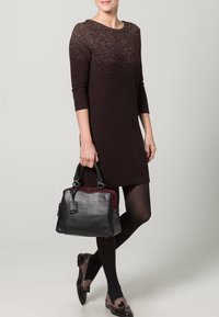 Sort læderhåndtaske med bordeaux detaljer. Glat tekstur, rektangulær form, dobbelte håndtag. Modellen bærer en mørk kjole og mønstrede sko.