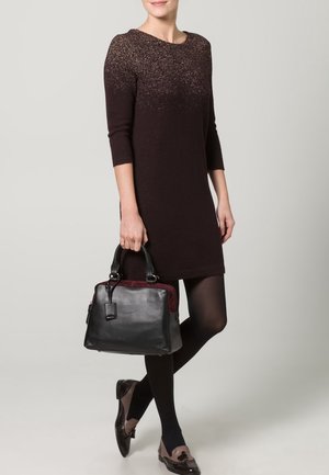 Czarna skórzana torebka z bordowym akcentem. Gładka faktura, prostokątny kształt, podwójne uchwyty. Model nosi ciemną sukienkę i wzorzyste buty.