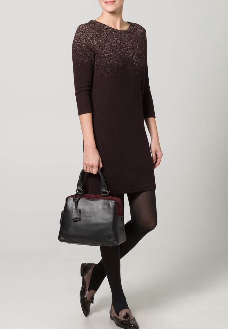 Sort læderhåndtaske med bordeaux detaljer. Glat tekstur, rektangulær form, dobbelte håndtag. Modellen bærer en mørk kjole og mønstrede sko.