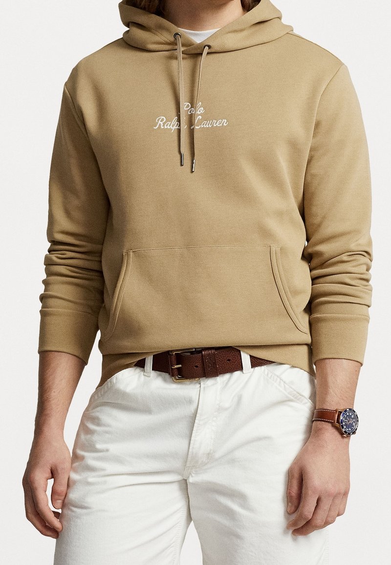 Homme portant un sweat à capuche beige avec un texte blanc, un pantalon blanc, une ceinture en cuir marron et une montre-bracelet en cuir marron avec un cadran bleu foncé.