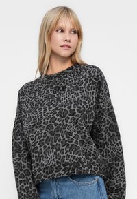 Donna bionda che indossa una felpa nera e grigia con stampa leopardata e logo ricamato, abbinata a jeans blu, mentre guarda di lato.