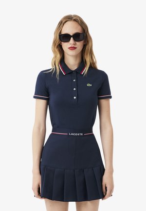 Donna che indossa un abito polo Lacoste blu navy con rifiniture bianche e rosse, gonna plissettata e occhiali da sole neri rettangolari su sfondo bianco.
