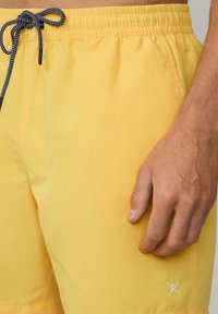 Žute kupaće šortse s elastičnim pojasom i užeom. Imaju mali bijeli logo sa strane i glatku, laganu tkaninu.