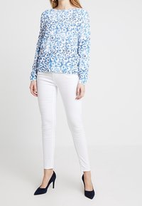 Blaues langärmliges Oberteil mit kreisförmigen Mustern, kombiniert mit weißen Slim-Fit-Hosen und navyblauen High Heels. Glatte Stofftextur, lässige Passform.