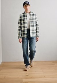 Chemise flanelle à carreaux verts et blancs, t-shirt blanc en dessous, jeans en denim bleu et baskets beiges avec des lacets gris. Fond en parquet.