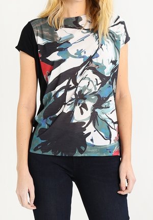 Femme portant un haut manches courtes noir avec un motif floral abstrait surdimensionné en blanc, bleu, sarcelle et rouge, associé à un pantalon foncé.