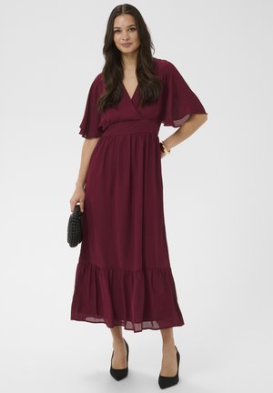 Femme portant une robe midi bordeaux avec des manches volantées, des talons noirs, tenant une pochette noire tissée, debout devant un fond uni.