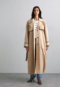 Beige trenchcoat met zwarte bies, grote kraag en knoopsluiting. Heeft lange mouwen en een relaxte pasvorm, gecombineerd met denim en laarzen.