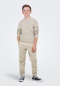 Beige crewneck sweatshirt met lange mouwen over een wit shirt, gecombineerd met beige sweatpants en zwarte sneakers. Eenvoudig ontwerp, casual pasvorm.