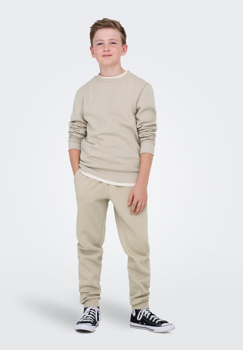 Beige crewneck sweatshirt met lange mouwen over een wit shirt, gecombineerd met beige sweatpants en zwarte sneakers. Eenvoudig ontwerp, casual pasvorm.