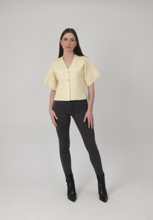 Kobieta z długimi ciemnymi włosami, ubrana w bluzkę na guziki w jasnożółtym kolorze, czarne obcisłe dżinsy oraz czarne botki, stojąca na tle jednolitego tła.