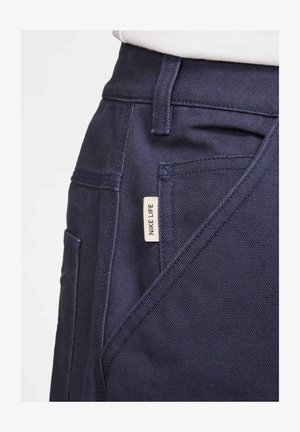 Marineblaue Hosen aus einem strukturierten Stoff, mit einer Vordertasche und einem kleinen, seitlich aufgenähten "NIKE LIFE"-Label.