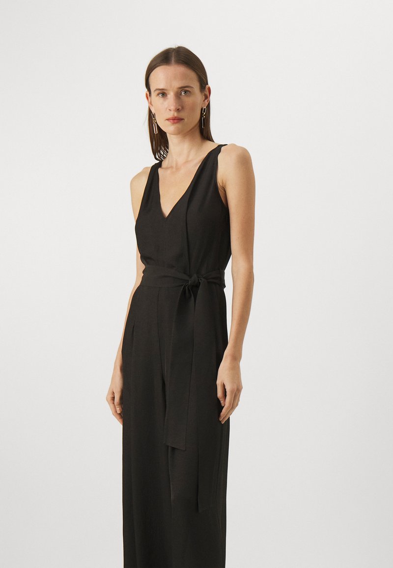 Svart ermeløs jumpsuit med dyp V-hals, tettsittende midje og bindebelte i stoff. Glatt tekstur, rett ben-design.
