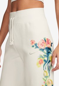 Närbild av vita joggingsbyxor med dragsko i midjan och färgglad blommönstring, med Nike-logotypen på höger lår.
