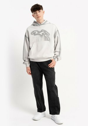 Młody mężczyzna w jasnoszarej bluzie z kapturem o abstrakcyjnym wzorze, w luźnych czarnych spodniach i białych trampkach, stojący na jednolitym białym tle.