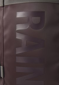 Brown waterproof backpack featuring a large, embossed "RAIN" text. Material appears smooth with a matte finish. Includes grey fabric accents.