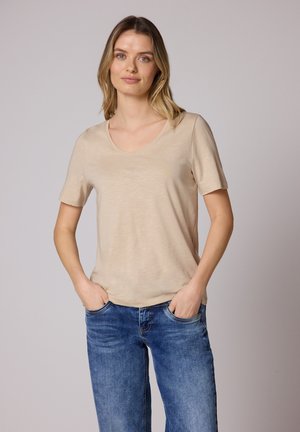 Kobieta o blond włosach, ubrana w beżową koszulę z krótkim rękawem i niebieskie jeansy, stojąca z rękami w kieszeniach na jednolitym tle.