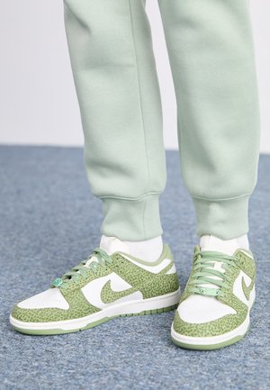 Fødder iført hvide og grønne Nike-sneakers med tekstureret mønster, kombineret med lysegrønne joggingbukser og hvide sokker på blåt tæppe.