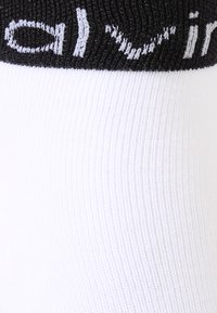 White cotton-blend socks with ribbed texture, featuring a black elastic cuff with white logo text prominently displayed.
