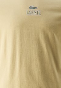 Złoto-beżowa koszulka z bawełny z małym zielonym logo krokodyla i tekstem "LACOSTE" w srebrnym kolorze. Gładki, lekki materiał.