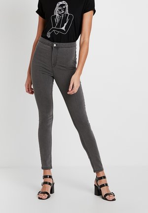 Pantalones vaqueros gris de talle alto y ajustados, fabricados en tejido de mezclilla, con cierre de botón y bolsillos frontales, combinados con una camiseta negra y sandalias de tacón.