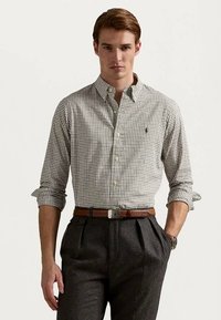 Νέος άνδρας που φοράει ένα καρό πουκάμισο με κουμπιά και ρολαρισμένα μανίκια, φορώντας σκούρες πτυχές παντελόνια, στέκεται με το ένα χέρι στην τσέπη.