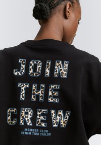 Svart sweatshirt med djärv text "GÅ MED I GÄNGET" i leopardmönster och blå kant, med mindre text nedanför. Mjuk tygkänsla.