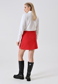 Frau mit glattem blonden Haar, die ein weißes Langarmoberteil, einen roten Minirock mit seitlichem Reißverschluss, goldene Ohrringe und schwarze kniehohe Stiefel trägt.