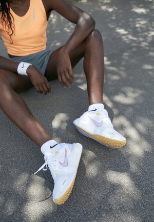 Coureur assis sur l'asphalte, portant des baskets blanches Nike avec des semelles en gomme, des chaussettes blanches Nike, un short gris et un débardeur orange.