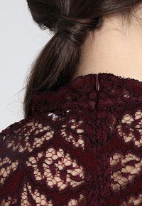 Burgundská krajková blůza s vysokým límcem a propracovanými květinovými vzory, s uzavíráním na knoflíky na zadní straně a texturovanou tkaninou.