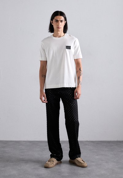White cotton t-shirt with a black patch pocket, paired with black patterned trousers and light brown slip-on shoes. Simple, modern design.