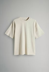 Gai krēmveida īsām piedurknēm t-krekliņš, kas izgatavots no mīkstas auduma. Tam ir apaļa kakla izgriezums un brīva piegriezuma forma ar pazeminātiem pleciem. Nav redzamu rakstu vai akcentu.