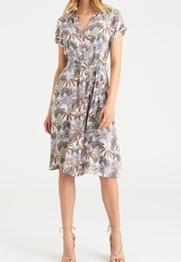 Kobieta ubrana w kwiecistą sukienkę z krótkim rękawem i paskiem, w liście i kwiaty, stojąca na neutralnym tle w beżowych sandałach na obcasie.