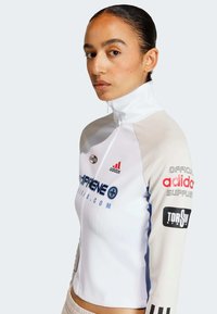Vit sportig zip-up tröja med långa ärmar. Innehåller svarta Adidas-strimmor, logotyper och blå grafisk text. Slät textur och figurnära design.