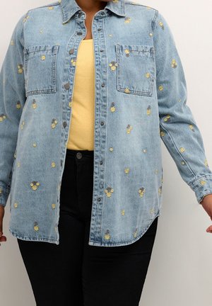 Personne portant une veste en denim bleu clair avec un imprimé floral jaune, par-dessus un haut jaune et un pantalon noir, sur un fond clair uni.