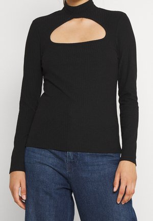 Femme portant un haut noir côtelé à manches longues avec une grande découpe courbe sur la poitrine, associé à un jean en denim bleu.