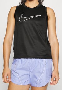 Černý sportovní top bez rukávů s síťovanou texturou a bílým logem Nike, doplněný o světle fialové šortky s bílými bočními akcenty.