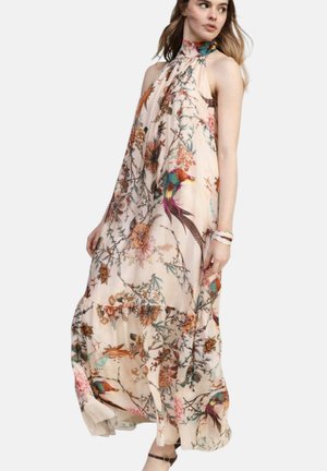 Donna indossa un abito floreale senza maniche lungo fino al pavimento con motivi di uccelli e rami, in piedi con un braccio rilassato e guarda di lato.