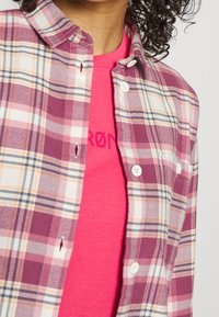 Personne portant un t-shirt rose sous une chemise à carreaux rose et blanche partiellement boutonnée, avec des cheveux bouclés foncés visibles en haut.