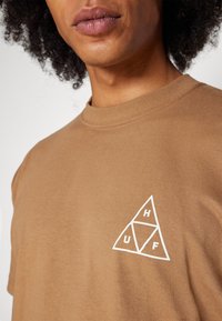 Camiseta de algodón marrón con un logo triangular blanco que presenta las letras "H", "U" y "F". Cuello redondo acanalado y mangas cortas. Textura suave.