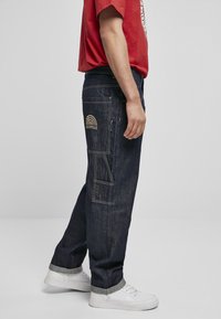 Pantaloni cargo din denim închis, cu multiple buzunare, având un logo curcubeu brodat cu fir auriu pe buzunarul din spate. Purtați cu sneakers albi.
