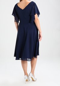 Vestido de gasa azul marino con mangas acampanadas, diseño de espalda en V y falda a la altura de la rodilla. Llevado con zapatos de tacón alto plateados.
