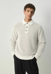Pulover alb tricotat cu un model texturat, mansete cu nervuri și guler alb; are o închidere cu trei nasturi, purtat peste pantaloni negri.